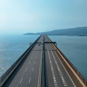 An aerial view of a long highway bridge stretching across the sparkling sea towards a distant, hilly coastline.