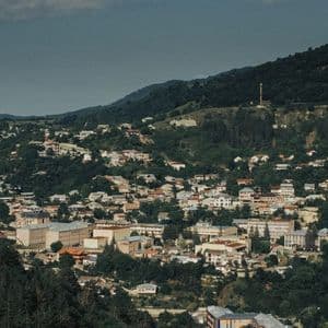 Une vue plongeante sur une ville avec des bâtiments dispersés sur une colline escarpée et boisée, sous un ciel partiellement nuageux.