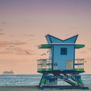 A blue and green lifeguard tower on Miami Beach at sunset, with a cruise ship on the ocean and seagulls flying by.