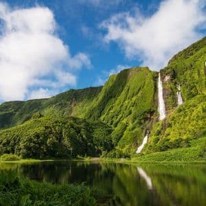 Múltiples cascadas caen por una exuberante ladera verde hacia un lago tranquilo, que refleja el cielo azul y las nubes.