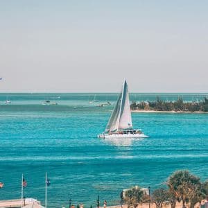 A WeRoad group trip sails on a white catamaran across turquoise water, viewed from a waterfront plaza under a clear sky.