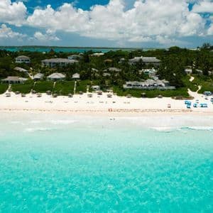 An aerial view of a tropical beach with white sand, clear turquoise water, and resorts nestled in lush green foliage under a partly cloudy sky.