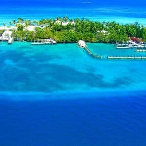 An aerial view of a small tropical island with palm trees and docks, surrounded by turquoise and deep blue ocean water.