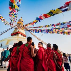 Un groupe de moines bouddhistes en robes rouges se tiennent près d'un stupa blanc, sous des drapeaux de prière colorés qui flottent dans un ciel bleu.