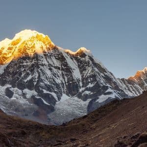 La luz dorada del amanecer o el atardecer ilumina los picos nevados de una cordillera, vistos desde un valle rocoso.