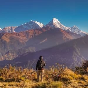 Ein Wanderer mit Rucksack steht auf einem grasbewachsenen Grat und blickt auf ein Panorama schneebedeckter Berge unter klarem Himmel.