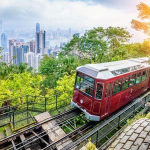 Un funicular rojo asciende por una vía en una exuberante ladera verde, con una vista panorámica de una moderna ciudad al fondo.