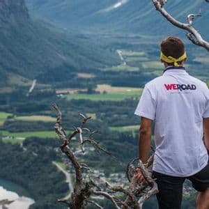 Un uomo con una maglietta WeRoad si trova su una scogliera, ammirando una vasta valle verde con un fiume e montagne.