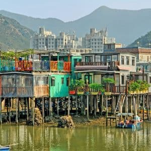 Coloridas casas sobre pilotes en un pueblo se alzan sobre el agua, con barcos amarrados cerca y montañas al fondo.