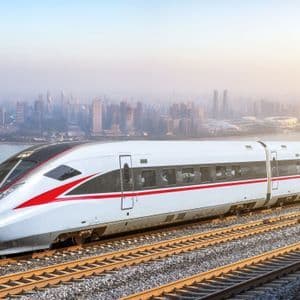 Un tren blanco de alta velocidad con una franja roja viaja por las vías, con vistas al horizonte de una ciudad moderna al otro lado del agua al amanecer.