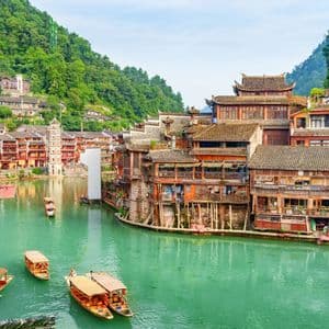 Un pueblo antiguo con casas tradicionales de madera sobre pilotes y botes en un río turquesa, rodeado de exuberantes montañas verdes.