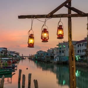 Tres farolillos rojos iluminados cuelgan sobre un canal en un pueblo palafítico al atardecer, con barcas amarradas a lo largo del agua.