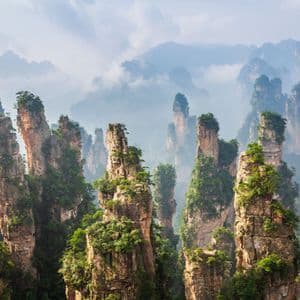 Imponentes formaciones rocosas cubiertas de vegetación verde se elevan desde un paisaje montañoso brumoso bajo un cielo nublado.