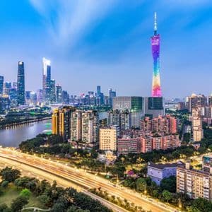 El horizonte de una ciudad moderna al anochecer, con un río, estelas de luces de una autopista y una torre destacada iluminada con los colores del arcoíris.