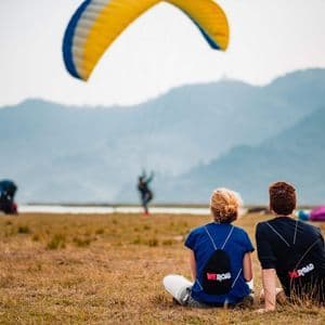 Deux participants d'un voyage de groupe WeRoad sont assis dans un champ herbeux, contemplant un parapente jaune et bleu au-dessus de montagnes lointaines.