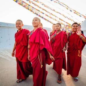 A group of young Buddhist nuns in red robes walking and smiling outdoors under colorful prayer flags next to a white stupa.