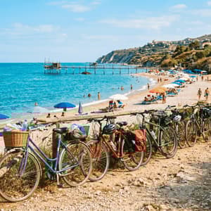 Una fila di biciclette parcheggiate su un sentiero costiero si affaccia su una spiaggia sabbiosa con acqua turchese e un pontile di legno.