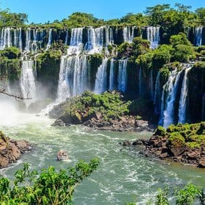 Una piccola barca turistica naviga sul fiume turbolento alla base di un'ampia cascata a più livelli, coperta da una lussureggiante vegetazione verde.