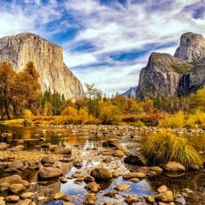 A wide valley with a rocky river, autumn foliage, and towering granite mountains under a partly cloudy sky.