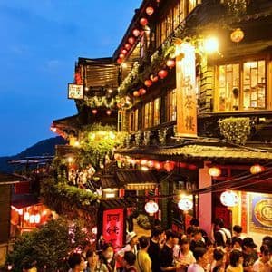 Une rue animée à flanc de colline au crépuscule, avec des bâtiments traditionnels à plusieurs étages ornés de lanternes en papier rouges lumineuses.