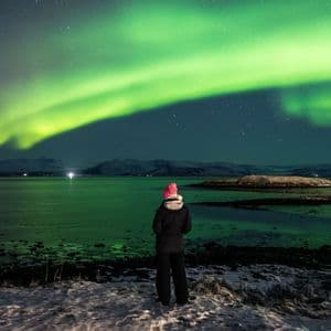 Una persona con un gorro rosa de pie en una orilla nevada observando la aurora boreal verde iluminar el cielo nocturno sobre una gran masa de agua.