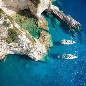 Una vista aerea di due barche a vela ancorate in acqua turchese accanto a scogliere rocciose bianche e una caletta appartata.