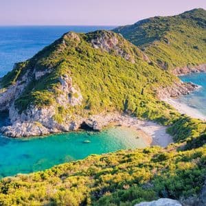 Ein Panoramablick auf eine üppig grüne Landzunge mit felsigen Klippen, die auf das türkisfarbene Meer treffen und einen abgelegenen Sandstrand in einer Bucht bilden.