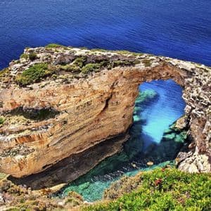 Ein natürlicher Steinbogen erstreckt sich von einer Klippe über türkisfarbenes und tiefblaues Meerwasser unter einem klaren Himmel.