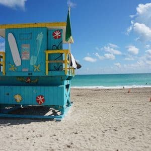 Una torre di salvataggio turchese e gialla si erge su una spiaggia sabbiosa, con l'oceano e un cielo azzurro parzialmente nuvoloso sullo sfondo.