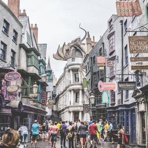 Una multitud de personas caminando por una calle empedrada flanqueada por edificios fantásticos, con un gran dragón posado en la azotea de un banco.