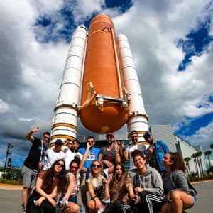 Un gruppo WeRoad in viaggio posa per una foto davanti a un serbatoio esterno di uno space shuttle e ai razzi propulsori, sotto un cielo nuvoloso.