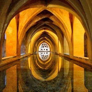 A long, arched hallway illuminated by golden light is reflected symmetrically in a narrow pool of still water.