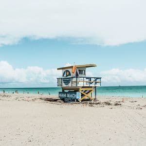 Ein bunter Rettungsschwimmerturm mit der Aufschrift 'Miami Beach' steht an einem Sandstrand mit dem Meer und einem bewölkten Himmel im Hintergrund.