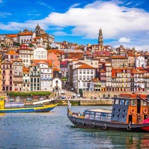 Two traditional boats sail on a river in front of a hillside city with colorful buildings under a partly cloudy blue sky.
