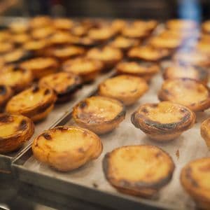 A close-up of freshly baked Portuguese egg tarts, Pastéis de Nata, with caramelized tops arranged on metal baking trays.