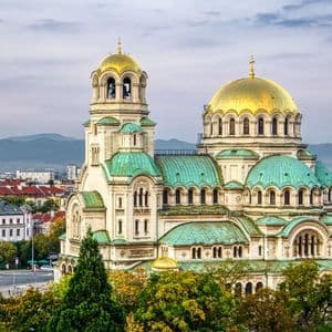 An ornate cathedral with large golden domes and teal roofs overlooks a cityscape with mountains in the background.