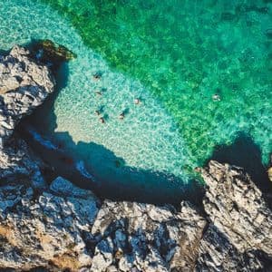 Una vista aerea di persone che nuotano nelle acque limpide e turchesi di una cala rocciosa.