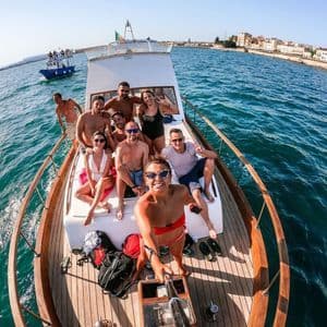 Une femme prend un selfie de groupe avec son voyage WeRoad sur le pont d'un bateau, avec la mer et une ville côtière derrière elle.