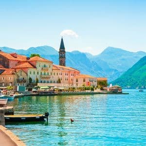 A coastal town with red-tiled roofs and a stone bell tower on the edge of a turquoise bay, with mountains in the distance.