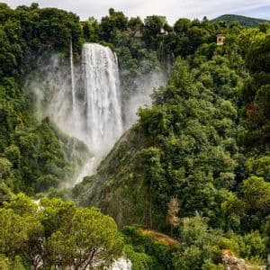 Una grande cascata scende da una scogliera rocciosa in una foresta lussureggiante e verde sotto un cielo nuvoloso.