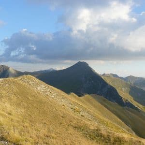 Uno stretto sentiero escursionistico corre lungo un crinale montuoso erboso, con altre cime visibili sullo sfondo sotto un cielo parzialmente nuvoloso.