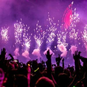 Des feux d'artifice explosent au-dessus d'une foule en silhouette lors d'un festival de musique avec des lumières violettes.