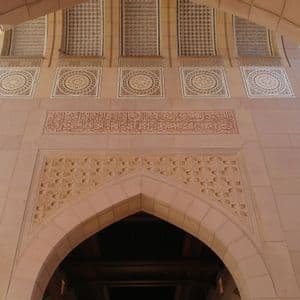 A low-angle view of an ornate building facade with geometric carvings, tile mosaics, and Arabic calligraphy, framed by large stone arches.