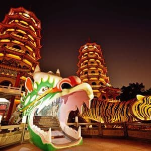 Deux grandes pagodes brillamment illuminées se découpent sur un ciel nocturne, avec d'imposantes statues colorées d'un dragon et d'un tigre au premier plan.