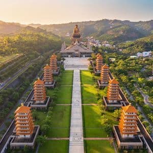 Vue aérienne d'un complexe de temples avec des pagodes bordant un chemin central menant à une grande statue de Bouddha, sur fond de collines verdoyantes.