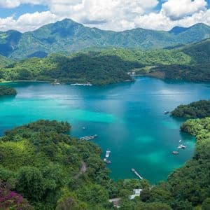 Vue aérienne d'un lac turquoise éclatant niché au cœur de montagnes verdoyantes, avec de petits bateaux parsemant l'eau sous un ciel nuageux.