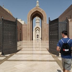 Un hombre con una camisa azul y una mochila WeRoad camina por una puerta ornamentada hacia el patio soleado de una mezquita con grandes arcos.