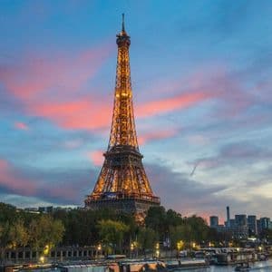 La Torre Eiffel illuminata si erge contro un cielo colorato al tramonto, con un fiume e lo skyline della città in primo piano.