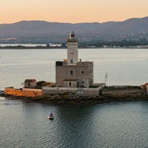 Un faro storico sorge su una piccola isola rocciosa in mezzo al mare, con una barchetta che galleggia nelle vicinanze sotto un cielo infuocato dal tramonto.