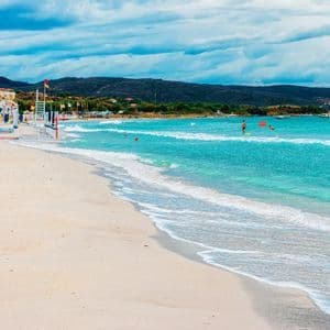 Una spiaggia di sabbia incontra l'oceano turchese, con piccole onde che lambiscono la riva. Sullo sfondo si scorgono lettini, edifici e colline.